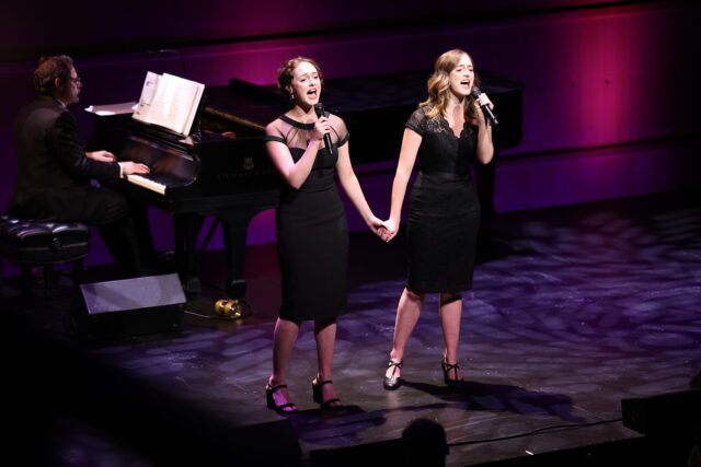 Two women hold hands and sing into microphones in front of a pianist