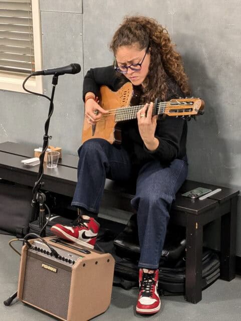 A young woman sits on a bench, playing flamenco guitar with one foot perched on an amp